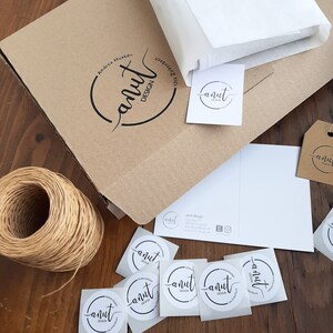 Puede incluir: Una caja de cart&oacute;n marr&oacute;n con el texto "anut DESIGN" y el nombre "Andrea Husken" impreso en ella. La caja est&aacute; rodeada de papel de seda blanco, una tarjeta de visita y varias pegatinas redondas con el texto "anut DESIGN".