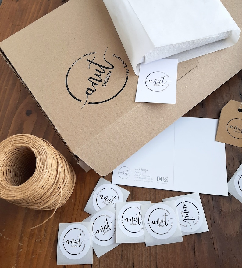 Puede incluir: Una caja de cart&oacute;n marr&oacute;n con el texto "anut DESIGN" y el nombre "Andrea Husken" impreso en ella. La caja est&aacute; rodeada de papel de seda blanco, una tarjeta de visita y varias pegatinas redondas con el texto "anut DESIGN".