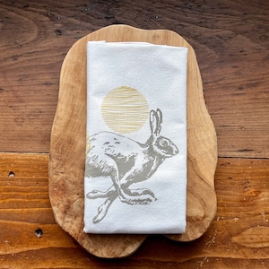 Könnte beinhalten: Ein weißes Geschirrtuch mit einer grauen Hasenillustration und einer gelben Sonnengrafik. Der Hase rennt und die Sonne ist im Hintergrund.