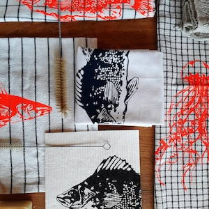 Puede incluir: Un juego de cuatro toallas de cocina con diferentes dise&ntilde;os de peces. Las toallas son blancas con peces negros y rojos impresos en ellas. Las toallas est&aacute;n dobladas y dispuestas sobre una superficie de madera.