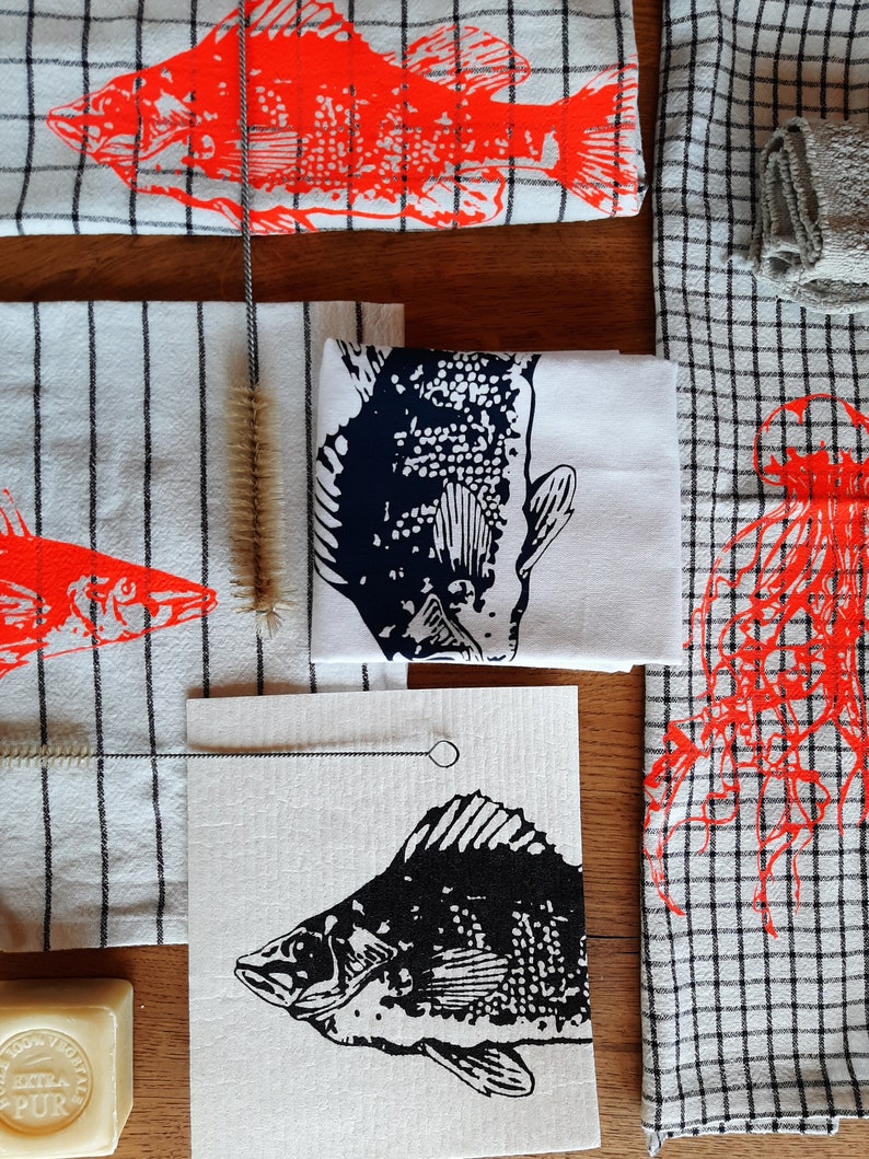 Puede incluir: Un juego de cuatro toallas de cocina con diferentes dise&ntilde;os de peces. Las toallas son blancas con peces negros y rojos impresos en ellas. Las toallas est&aacute;n dobladas y dispuestas sobre una superficie de madera.