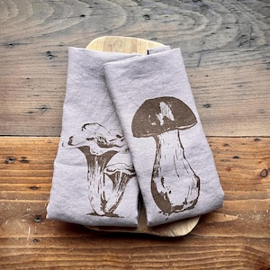 Op de afbeelding: Twee linnen theedoeken met een bruine paddenstoelenprint. Een theedoek heeft een enkele paddenstoel met een grote dop en een dikke steel. De andere theedoek heeft twee paddenstoelen met een delicate, waaiervormige dop.