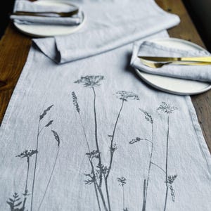 Op de afbeelding: Een lichtgrijze linnen tafelrunner met een zwart bloemenontwerp van wilde bloemen en grassen. De runner ligt op een houten tafel.
