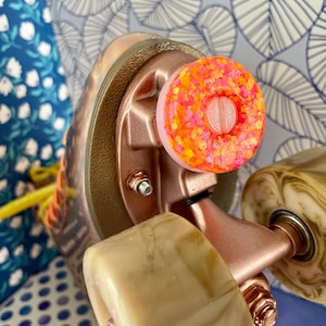 May include: Close-up of a roller skate with a pink and orange glitter wheel stopper. The skate has a rose gold frame and wheels with a marbled pattern.
