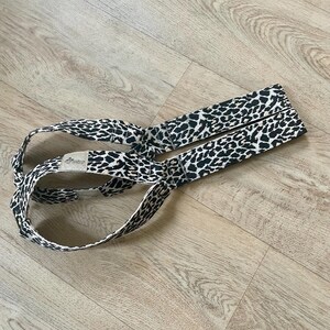 Peut inclure: Une sangle de yoga à imprimé léopard, avec un motif tacheté noir et blanc. La sangle est fabriquée dans un tissu durable et comporte une boucle à chaque extrémité. La sangle est conçue pour aider à l'étirement et aux postures.