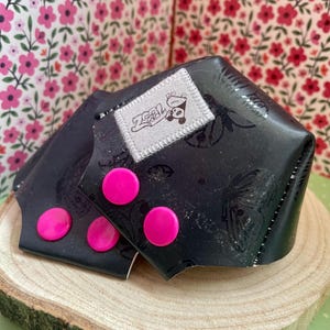 May include: Black wrist cuffs with a butterfly pattern and three hot pink buttons. Each cuff has a small white tag with a panda bear logo. The cuffs are displayed on a wooden slice.