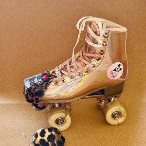 May include: A rose gold roller skate with matching laces and wheels. The skate features a leopard print toe cap with red star accents and a cartoon character sticker. A matching leopard print toe cap cover is also present.