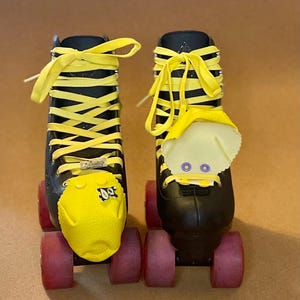 May include: Pair of black roller skates with yellow laces and toe stops. The skates have red wheels and a yellow protective cover on the toe. The laces are tied in a crisscross pattern.