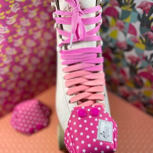 Puede incluir: Patín blanco con cordones rosas y un protector de dedos de color rosa y blanco con estampado de estrellas. El protector de dedos tiene dos botones rosas.
