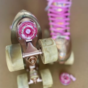 May include: Rose gold roller skates with pink laces and wheels. The skates have a metallic finish and are set against a neutral background. The wheels are a light, translucent colour.