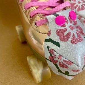 May include: Close-up of a rose gold roller skate with pink laces. A decorative mask with a floral design in pink glitter and green accents is attached to the laces. The skate is resting on a light-colored surface.