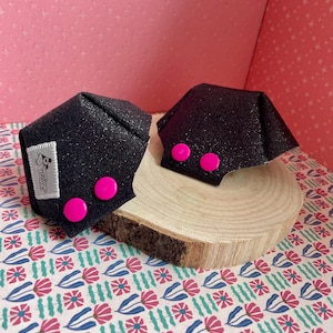 Puede incluir: Dos objetos negros brillantes estilo origami con detalles circulares de color rosa brillante. Uno tiene una pequeña etiqueta blanca con texto negro. Los objetos se exhiben sobre una rodaja de madera, sobre un fondo estampado con diseños florales rosas y azules.
