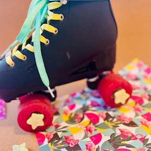 May include: Black roller skate with mint green laces and yellow eyelets. The red wheels have star-shaped accents. The skate rests on a colorful patterned surface with flamingo and geometric designs. Several star-shaped wax melts are also present.