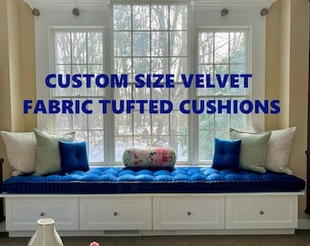 May include: A custom size velvet fabric tufted cushion in blue with white piping. The cushion is on a window seat with white drawers. The window seat is in front of a large window with a view of trees.
