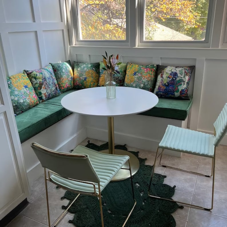 May include: A white round table with a gold base sits in a corner nook with a green velvet bench and two light green chairs with gold frames. The bench has floral patterned cushions in shades of yellow, green, and blue. A vase with white flowers sits on the table.