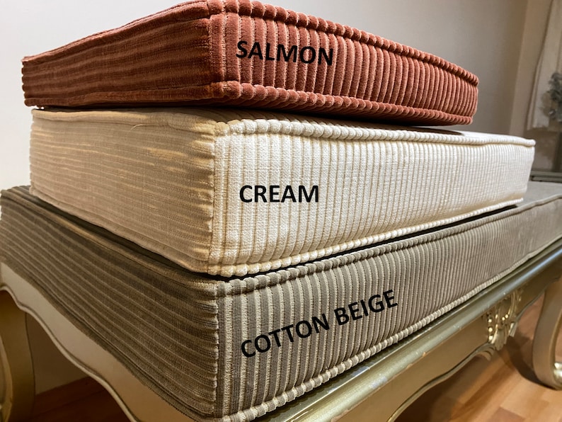 May include: Three corduroy cushion covers stacked on top of each other. The top cover is salmon, the middle cover is cream, and the bottom cover is cotton beige.
