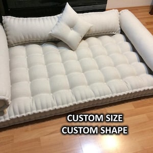 May include: A custom-sized, off-white tufted pet bed with bolster pillows and a square pillow. The bed has a rectangular shape with rounded corners and a decorative border. The text "CUSTOM SIZE CUSTOM SHAPE" is visible.