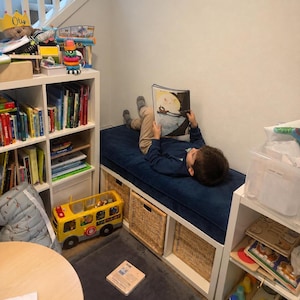 May include: A white storage bench with a dark blue cushion, featuring three woven wicker baskets underneath. A child lies on the bench reading an open book. The bench is positioned between two white shelving units filled with books and various toys.