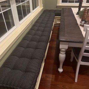 May include: A gray tufted bench cushion with a window behind it. The bench cushion is in front of a wooden table with a white base and a dark wood top.