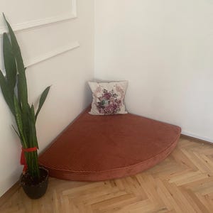 May include: A rust-coloured corner bench with a corduroy-like texture. A floral patterned cushion rests on the bench. A potted snake plant with green leaves and a red ribbon is in the corner. The bench is on a wooden floor.
