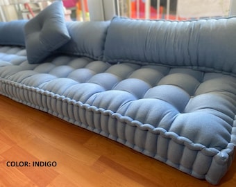 Puede incluir: Un cojín de suelo capitoné en azul índigo con una base rectangular gruesa con borde ribeteado y un cojín de respaldo largo a juego. La superficie tiene una cuadrícula de hendiduras capitoné profundas. El texto COLOR: INDIGO es visible.