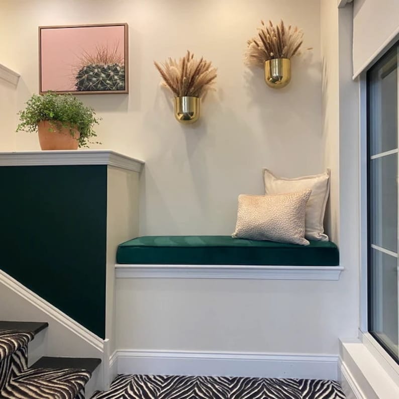 May include: A green velvet bench with two beige cushions sits in a corner of a room with white walls and a green accent wall. The bench is in front of a window with white blinds. Two gold planters with dried pampas grass hang on the wall above the bench. A framed print of a cactus hangs on the wall to the left of the bench.
