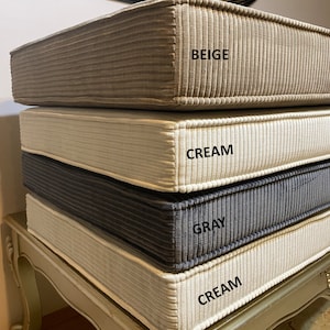 May include: Four stackable cushions in different colors. The top cushion is beige, the second is cream, the third is gray, and the bottom cushion is cream. The cushions are made of a textured fabric.