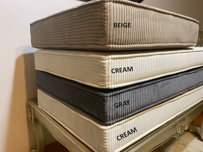 May include: Four stackable cushions in different colors. The top cushion is beige, the second is cream, the third is gray, and the bottom cushion is cream. The cushions are made of a textured fabric.