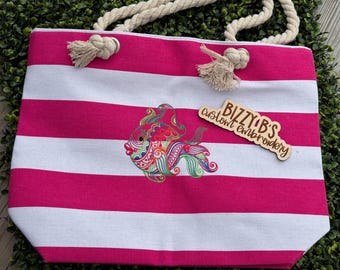 May include: A tote bag featuring horizontal hot pink and white stripes with thick cream-colored rope handles. The center displays a colorful embroidered fish with intricate patterns. A wooden sign reads BIZZY B'S custom embroidery.
