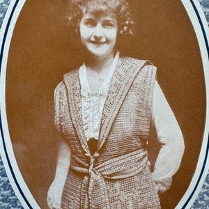 May include: Sepia-toned portrait of a woman wearing a crocheted vest with a tie at the waist. The vest is over a white blouse with a ruffled neckline. The image is framed within an oval border.