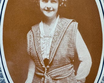 May include: Sepia-toned portrait of a woman wearing a crocheted vest with a tie at the waist. The vest is over a white blouse with a ruffled neckline. The image is framed within an oval border.