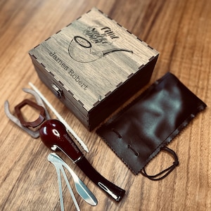 Może przedstawiać: Drewniane pudełko z wygrawerowanym tekstem "Fine Smoke Since" i "James Robert". Pudełko zawiera fajkę, narzędzia do fajki i zestaw do czyszczenia. Fajka jest czerwono-czarna i ma wygrawerowany tekst "James Robert". Pudełko stoi na drewnianej powierzchni.