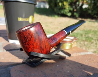 Pode incluir: Um cachimbo de madeira vermelho e marrom com um bocal preto. O cachimbo está apoiado em um suporte de couro preto.