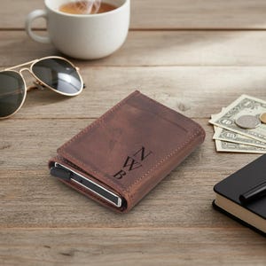 Könnte beinhalten: Ein braunes Lederportemonnaie mit den Initialen "NWB" auf der Vorderseite. Das Portemonnaie liegt auf einer Holzoberfläche, neben einer Sonnenbrille, einer Tasse Kaffee und etwas Bargeld.