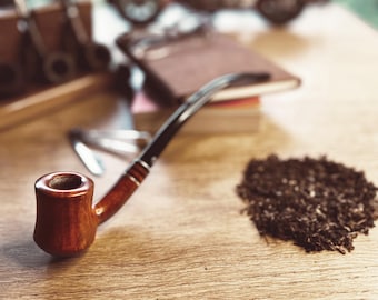 May include: A brown wooden pipe with a black stem and a bowl filled with dark brown tobacco. The pipe is sitting on a wooden table.