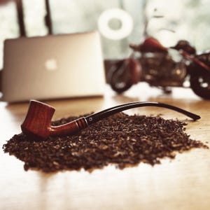 Könnte beinhalten: Eine braune und schwarze Holzpfeife mit einem schwarzen Schaft, die auf einem Haufen loser brauner Tabakblätter liegt.