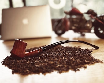 Pode incluir: Um cachimbo de madeira marrom e preto com um caule preto descansando sobre um monte de folhas de tabaco marrons soltas.