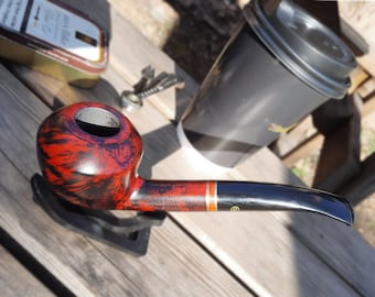 May include: A red and black tobacco pipe with a black stem and a gold band. The pipe is sitting on a wooden surface.