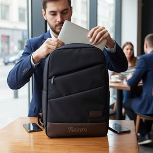 Può includere: Zaino nero con pannello inferiore marrone. Lo zaino ha una tasca con cerniera e una piccola toppa con il logo. Il nome "Aaron" è stampato sul pannello marrone. Una persona sta mettendo un laptop nello zaino.