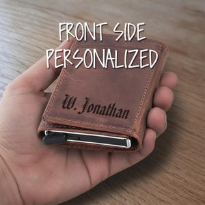 Könnte beinhalten: Braunes Lederportemonnaie mit dem Text "FRONT SIDE PERSONALIZED" und dem Namen "W. Jonathan" auf der Vorderseite eingraviert. Das Portemonnaie hat einen Metallverschluss und Kartenfächer. Das Portemonnaie wird in einer Hand gehalten.