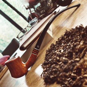 Könnte beinhalten: Eine braune Holzpfeife mit einem schwarzen Schaft und einem Kopf aus dunklem Holz. Die Pfeife liegt auf einem Buch und einem Haufen Tabak.