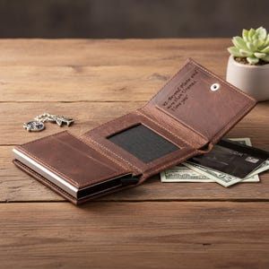 May include: Open brown leather wallet with a silver metal card holder. The wallet is open, revealing a black mesh pocket, credit cards, and cash. The inside of the wallet has the text "42 - Beyond Pluto and more than Uranus. I love you."