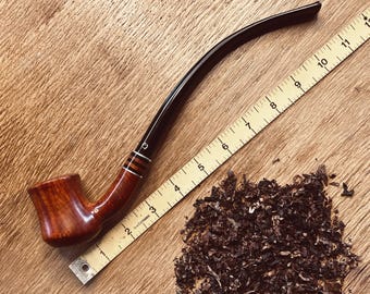 May include: A brown wooden pipe with a black stem and a brown and black band. The pipe is sitting on a wooden surface next to a pile of loose tobacco and a measuring tape.