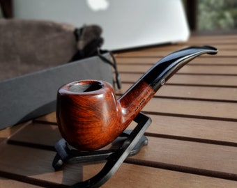 Pode incluir: Um cachimbo de tabaco de madeira polida, com um bocal escuro e curvo. A tigela é castanho-avermelhada, e o caule é preto. O cachimbo repousa sobre um suporte preto, mostrando o seu artesanato. Um acessório clássico.