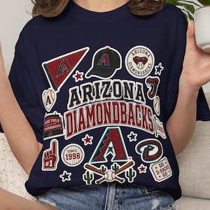 以下が含まれることがあります： ネイビーブルーのTシャツ。アリゾナ・ダイヤモンドバックスの野球チームの記念品をモチーフにしたグラフィックプリントが施されており、ペナント、野球帽、フォームフィンガー、バット、そして1998年と記されたArizona Diamondbacksの文字が描かれています。