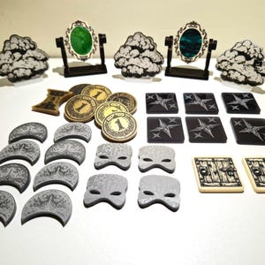 Può includere: Una collezione di componenti per giochi da tavolo che include tre gettoni nuvola in bianco e nero, due piccoli specchi incorniciati con inserti verdi e blu, monete color oro, tessere quadrate nere con disegni a stella, gettoni a mezzaluna grigi, gettoni maschera grigi e due tessere rettangolari con illustrazioni di porte.