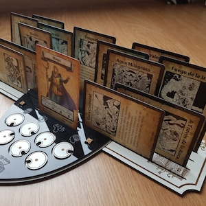 Könnte beinhalten: Ein schwarz-weißer Spielkartenhalter mit mehreren Karten mit Illustrationen und Text. Die zentrale Karte zeigt einen Charakter mit der Bezeichnung "Mago". Der Halter hat ein einzigartiges Design mit kreisförmigen Aussparungen.