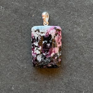Könnte beinhalten: Rechteckiger Anhänger mit silberner Öse. Der Anhänger zeigt einen farbenfrohen Stein mit einer Mischung aus schwarzen, weißen und rosa Farbtönen. Der Stein hat eine polierte Oberfläche vor einem dunklen Hintergrund.