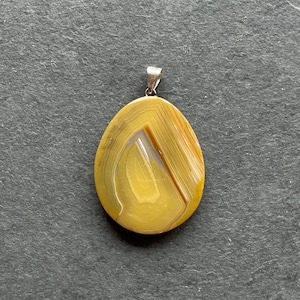 Könnte beinhalten: Ein gelber Achat-Anhänger mit einer silbernen Öse. Der Achat hat ein wirbelndes Muster aus Gelb und Weiß.