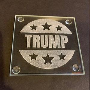 May include: A glass coaster with a white design of a circle with five stars and the word "TRUMP" inside. The coaster has four rubber feet on the bottom.